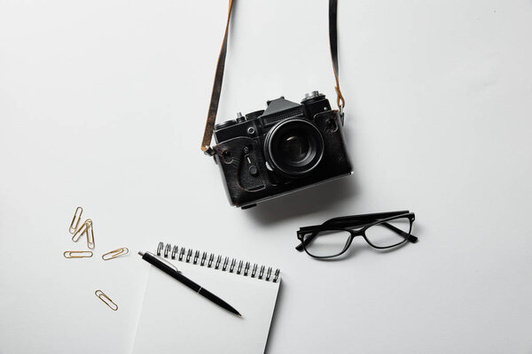 top view of photo camera, glasses, notepad with pen and stationery on white surface