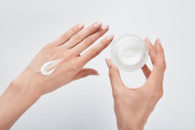 cropped view of woman applying cream on skin and holding jar isolated on white clipart