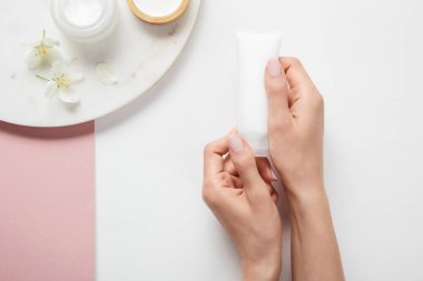 beyaz pembe yüzeyüzerinde kozmetik ve çiçek ile plaka yakınında krem tüp tutan kadın kırpılmış görünümü