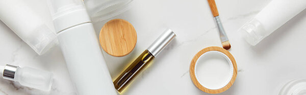 панорамный снимок кремовых трубок, банок для косметики, диспенсера, стеклянных и тушью бутылок с кистью для глаз на белой поверхности
