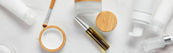 панорамный снимок кремовых трубок, банок для косметики, диспенсера, стеклянных и тушью бутылок с кистью для глаз на белой поверхности
