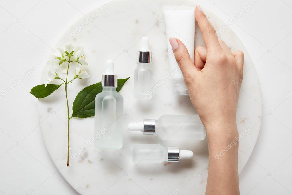 cropped view of woman hand with cream tube over plate with jasmine and cosmetic glass bottles on white surface