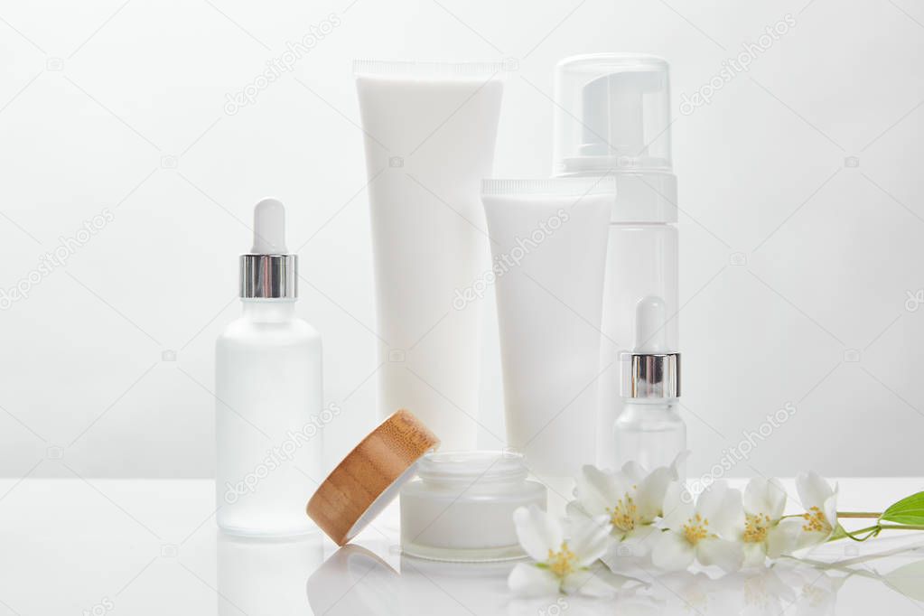jasmine flowers on white surface near glass bottles, cream in tubes, jar and cosmetic dispenser