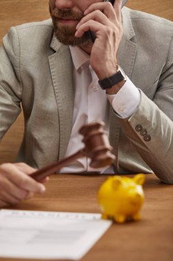 cropped view of businessman in suit talking on smartphone while holding wooden gavel near yellow piggy bank clipart