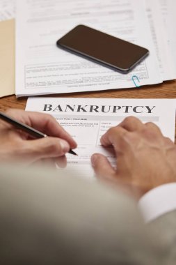 cropped view of businessman filling in bankruptcy form at wooden table with smartphone clipart