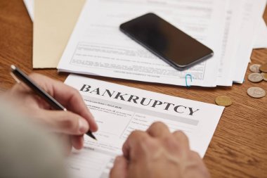 partial view of businessman filling in bankruptcy form at wooden table with smartphone clipart