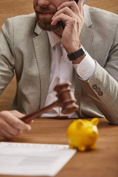黄色の貯金箱の近くに木製のガゼルを保持しながら スマートフォンで話すスーツを着たビジネスマンのトリミングビュー — ストック写真