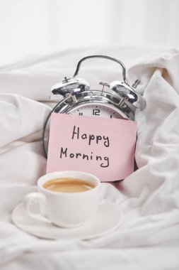 coffee in white cup on saucer near silver alarm clock with happy morning lettering on sticky note in bed clipart