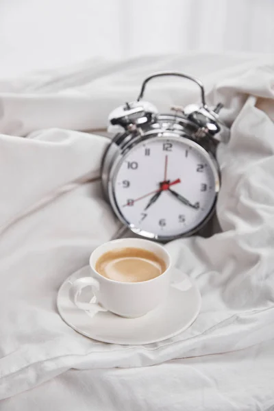 白い寝具の銀の目覚まし時計の近くに受け皿の白いカップでコーヒー — ストック写真