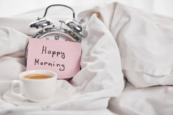咖啡在白色杯附近的银色闹钟与快乐的早晨信在床上的粘滞便笺 — 图库照片