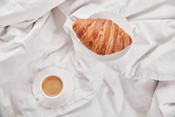 新鲜羊角面包的顶视图在盘上接近咖啡在白杯在床上的碟 — 图库照片