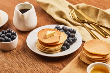 plate with pancakes and blueberries, jug with syrup near linen cloth with golden cutlery on wooden surface  clipart