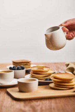 cropped view of man holding jug with syrup upon plate with pancakes and blueberries near cup of coffee and bowl with honey isolated on grey clipart