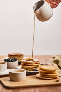 cropped view of man pouring tasty pancakes with syrup from jug near bowls with blueberries and honey, and cup of coffee isolated on grey clipart