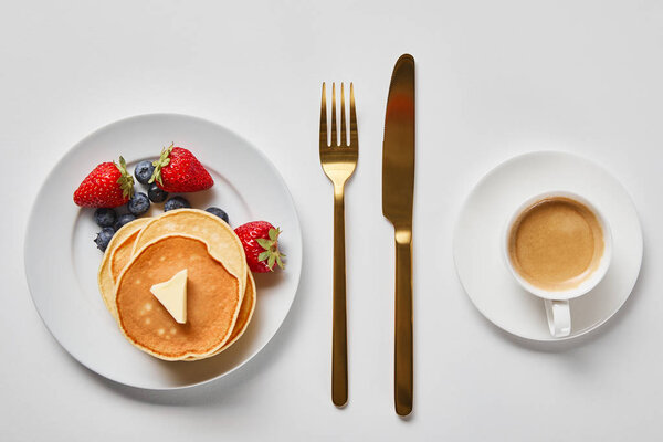 top view of tasty breakfast with pancakes, berries and cup of coffee near golden cutlery