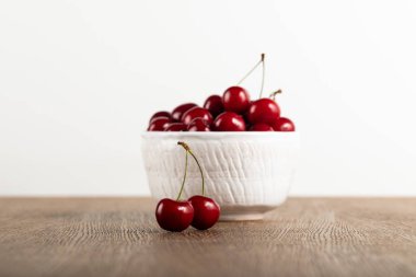 selective focus of red delicious cherries in white bowl and on wooden table isolated on white clipart