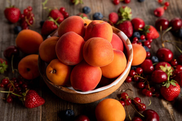 close up view of ripe delicious seasonal berries scattered around bowl with apricots on wooden table 