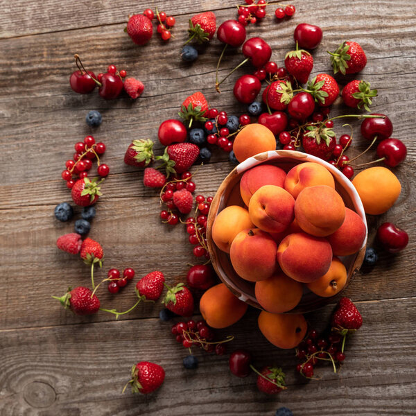 top view of ripe delicious seasonal berries scattered around bowl with fresh apricots on wooden table 