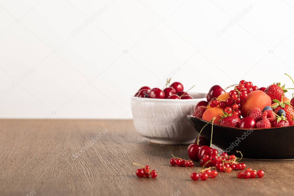 selective focus of cherries in white bowl and mixed berries on plate on wooden table isolated on white