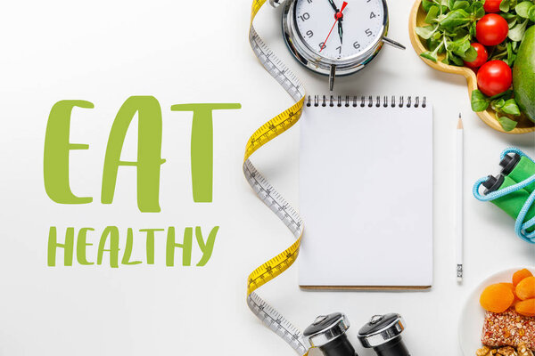 top view of sport equipment, measuring tape, alarm clock and diet food near empty notebook on white background with eat healthy lettering