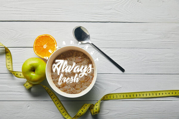 top view of measuring tape, spoon and breakfast cereal in bowl near apple and orange on wooden white background with breakfast always fresh lettering