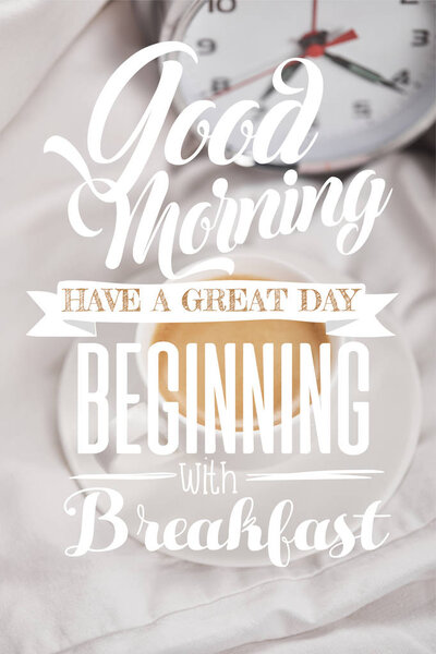 top view of coffee in white cup on saucer near silver alarm clock on bedding with good morning, have a great day beginning with breakfast lettering