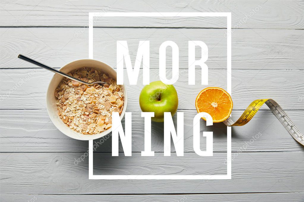 flat lay with breakfast cereal in bowl, apple, orange and measuring tape on wooden white background with morning lettering