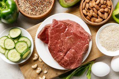 top view of raw meat on wooden chopping board near nuts, groats, eggs and green vegetables, ketogenic diet menu clipart