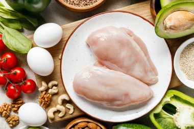top view of fresh raw chicken breasts on white plate near nuts, eggs and vegetables, ketogenic diet menu clipart