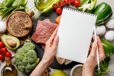 cropped view of woman holding empty notebook above food for ketogenic diet menu clipart