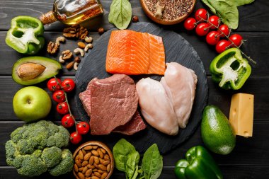 top view of raw salmon, meat and chicken breasts on wooden black table with vegetables and nuts, ketogenic diet menu clipart