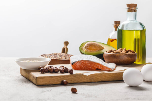 Свежий сырой лосось, орехи, яйца и авокадо на деревянной доске возле масла, выделенного на сером, кетогенном меню диеты
