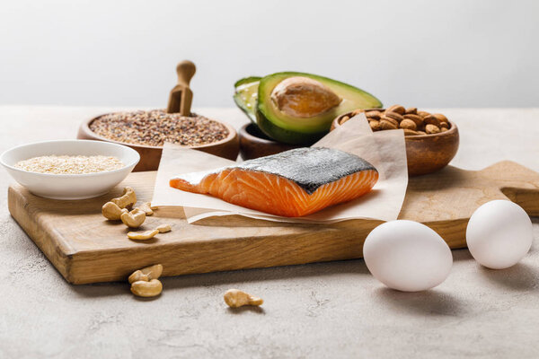 fresh salmon, nuts, eggs and avocado on wooden chopping board near oil isolated on grey, ketogenic diet menu