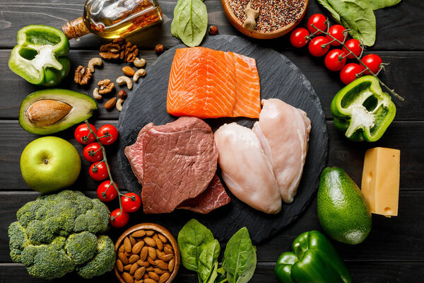 top view of raw salmon, meat and chicken breasts on wooden black table with vegetables and nuts, ketogenic diet menu