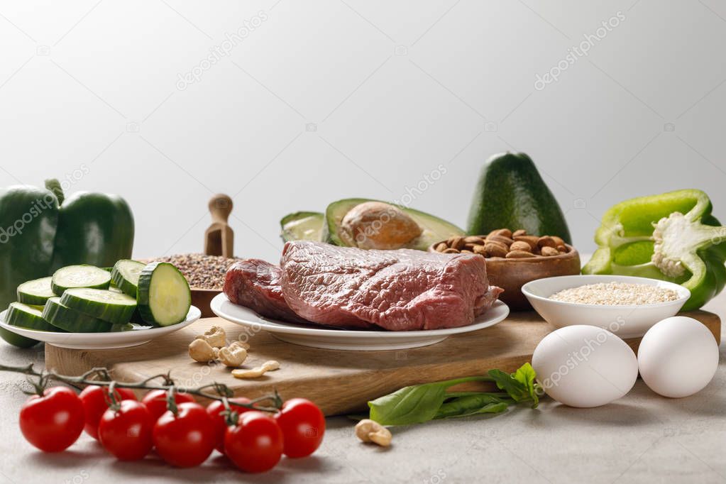 fresh raw meat on wooden chopping board near nuts and vegetables isolated on grey, ketogenic diet menu