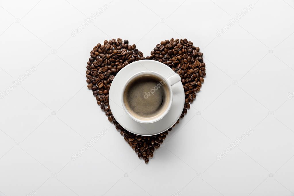 top view of tasty coffee in cup on saucer with heart made of coffee grains on white background