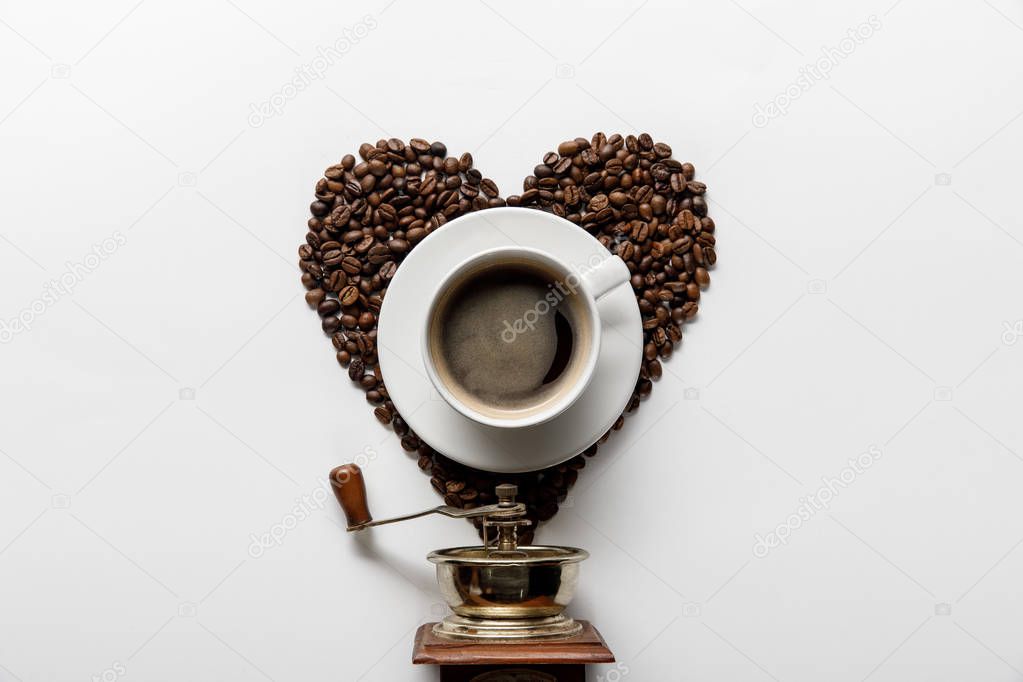 top view of coffee in cup on saucer with heart made of coffee grains near vintage coffee grinder on white background