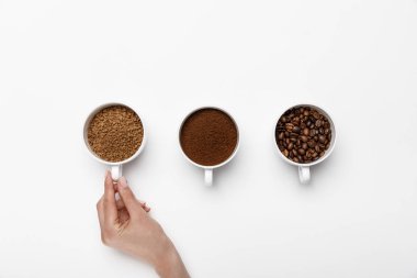 cropped view of female hand near three types of coffee grinding in cups on white background