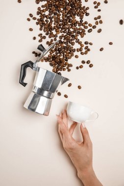 partial view of woman holding cup near aluminium coffee pot and scattered fresh coffee beans on beige background clipart
