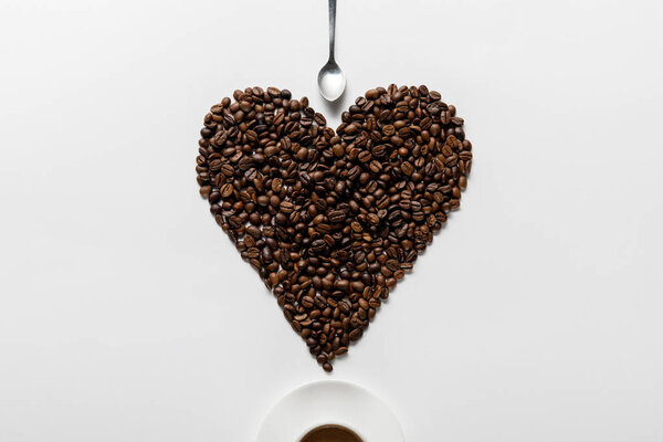 top view of coffee in cup on saucer with heart made of coffee grains near spoon on white background
