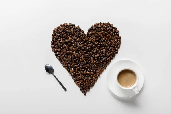 咖啡在杯在碟子上与心脏由咖啡谷物和勺子在白色背景的顶视图 — 图库照片