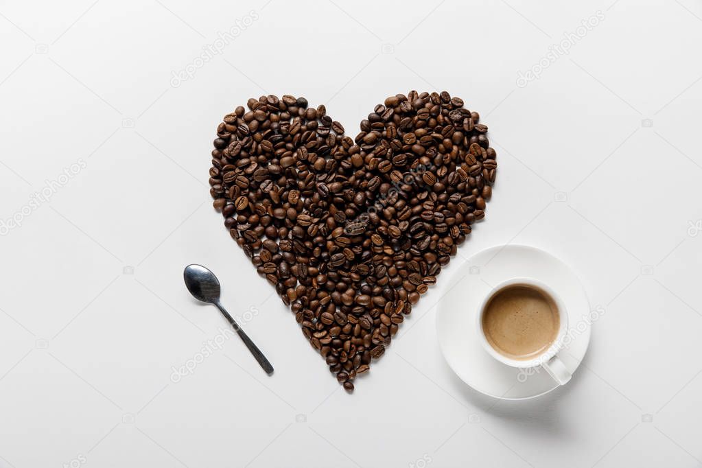 top view of coffee in cup on saucer with heart made of coffee grains and spoon on white background