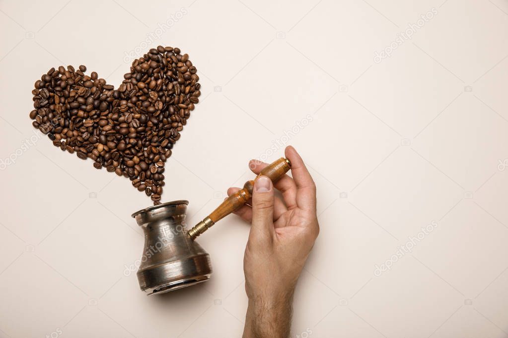 cropped view of man holding cezve near heart made of coffee grains on beige background