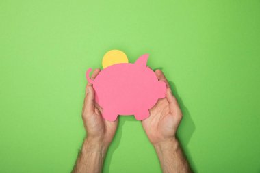 cropped view of man holding paper piggy bank with coin on green clipart