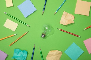top view of light bulb near scattered pencils and crumpled paper on green clipart
