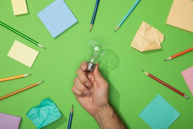 cropped view of man holding light bulb near pencils and paper on green clipart