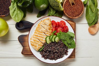 Ahşap doğrama tahtası üzerinde beyaz tabakta ızgara tavuk göğsü ve dilimlenmiş sebze ile pişmiş Quinoa üst görünümü