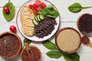 top view of cooked quinoa with grilled chicken breast and vegetables on white plate on wooden table with spinach leaves and raw seeds clipart