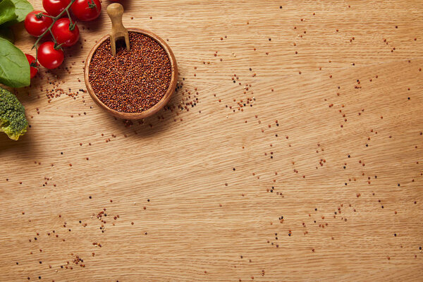 сверху вид на семена красного киноа в деревянной чаше рядом с красными помидорами, брокколи и листьями шпината на деревянном столе
