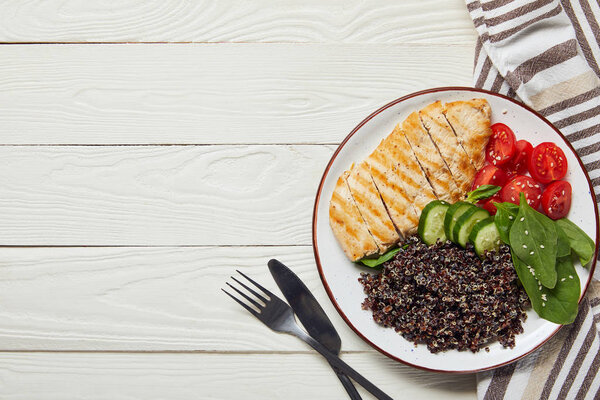 top view of cooked quinoa with grilled chicken breast and vegetables on white wooden table with cutlery and napkin